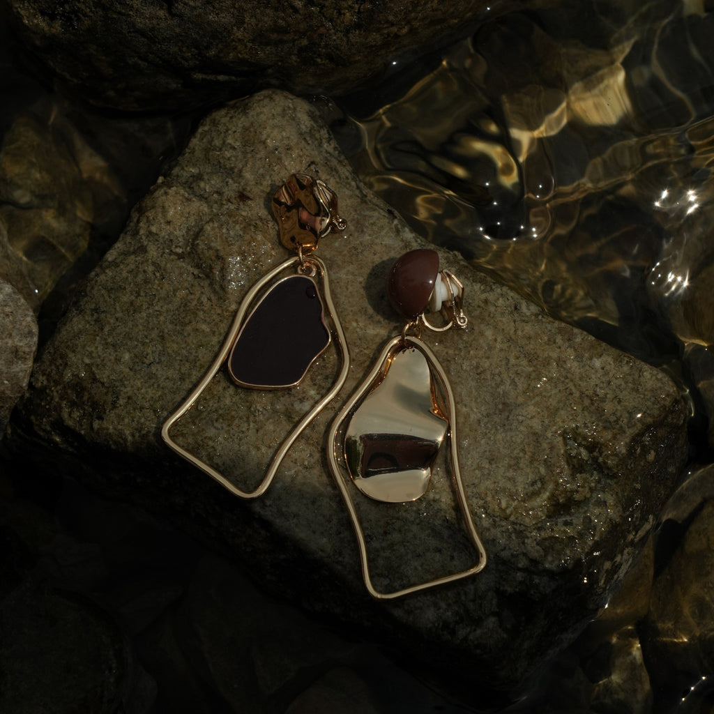 A pair of gold-tone statement earrings resting on wet rocks in shallow water. One earring features a dark, irregularly shaped stone, and the other features a polished metallic gold disc with a small brown bead accent.